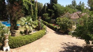 Un chemin mène à un espace piscine entouré d’une végétation luxuriante et à un bâtiment traditionnel doté d’un toit en tuiles.