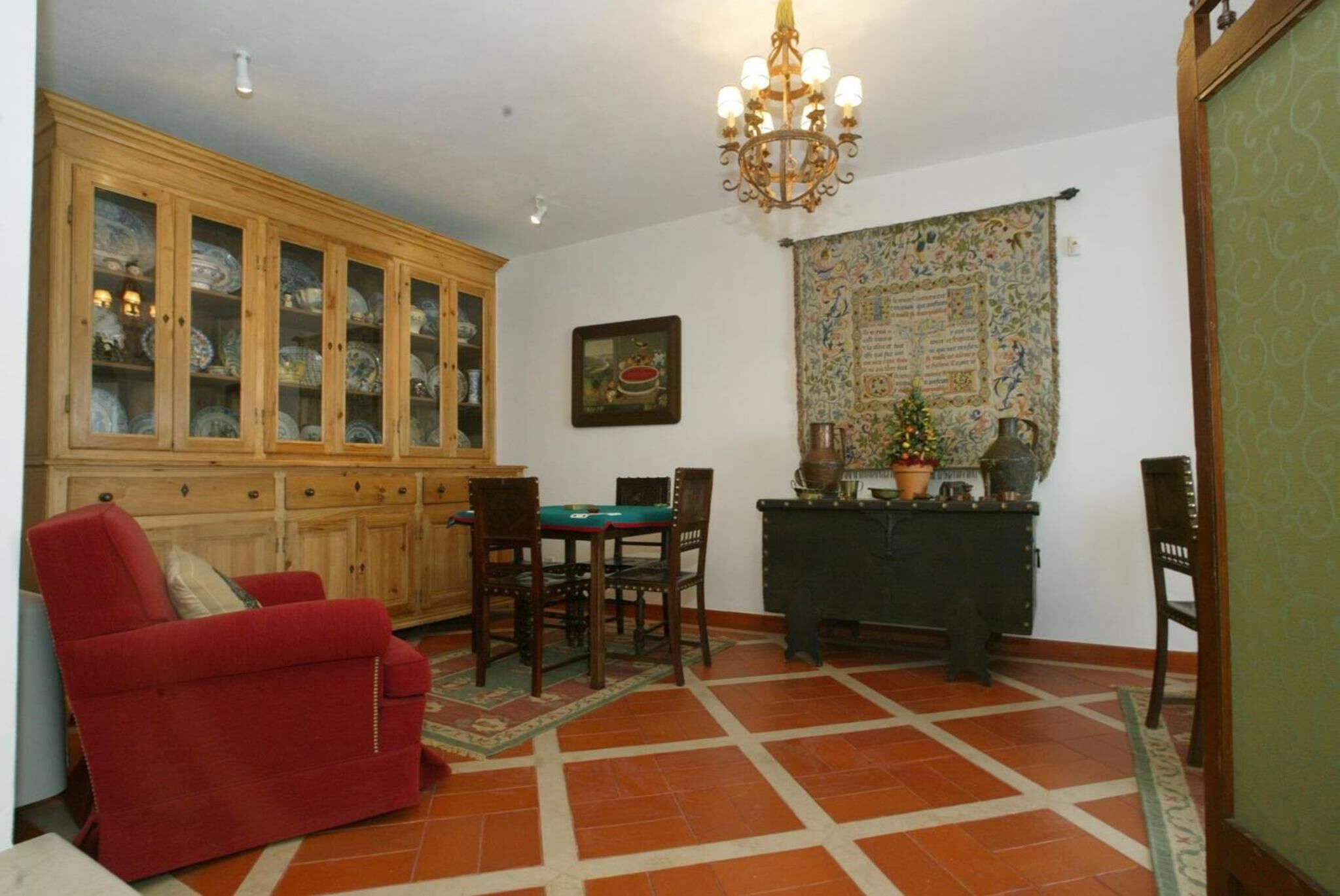 Una sala de estar con un sofá rojo, un aparador de madera con puertas de vidrio, una mesa de comedor con sillas, una araña y un tapiz en la pared.