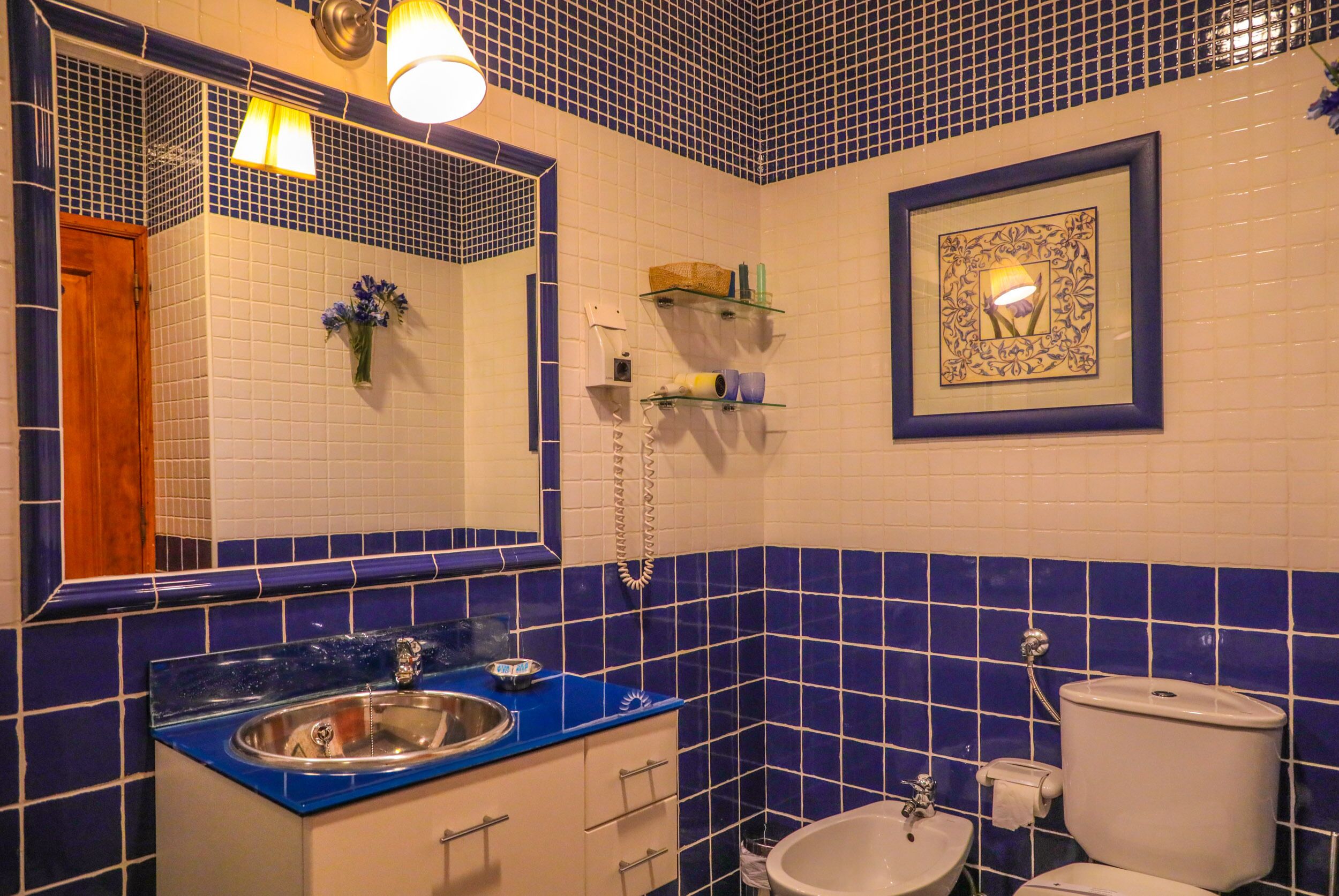 A bathroom with a blue-tiled wall, a sink with a blue countertop, a mirror, a toilet, and shelves with toiletries.