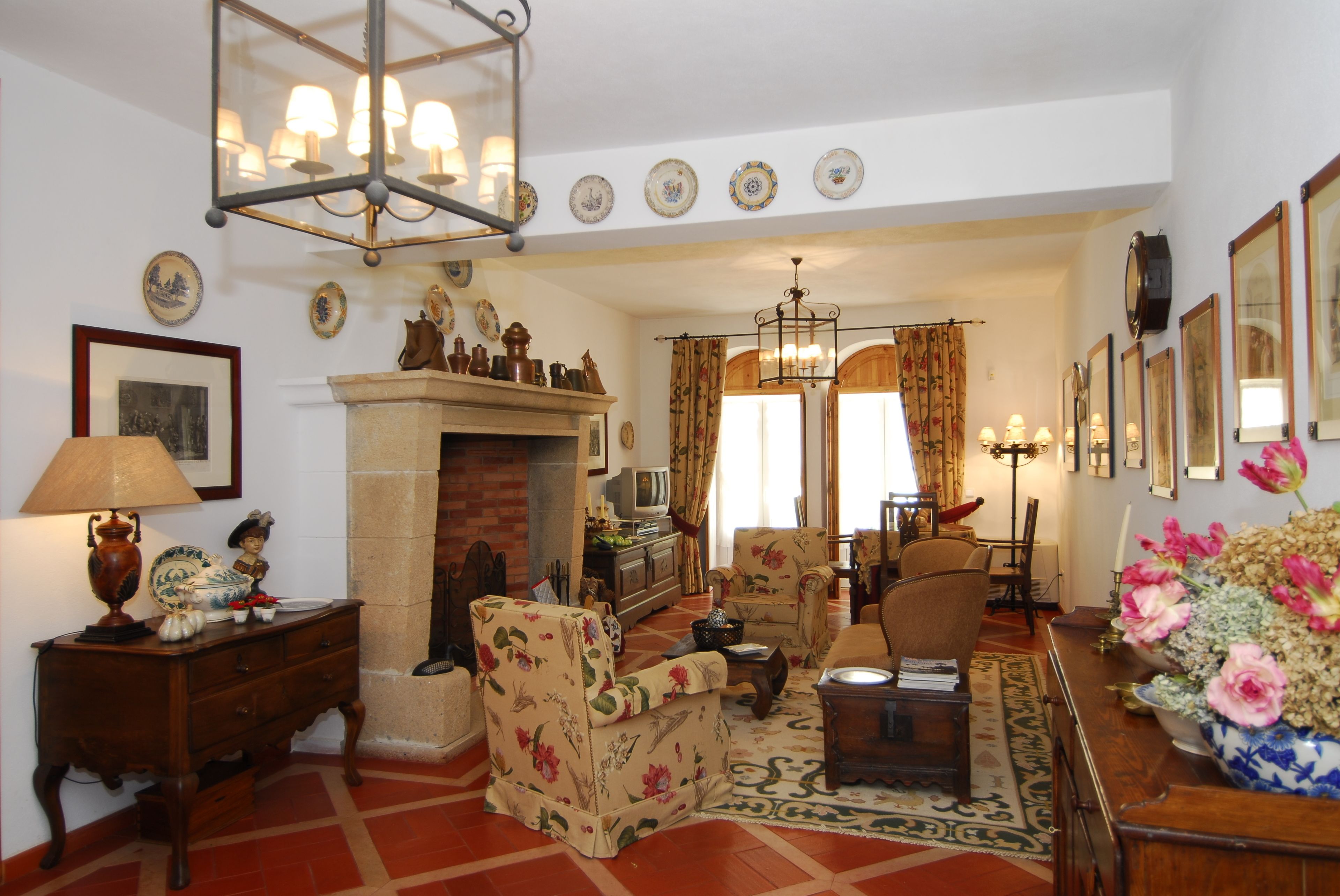 A traditional living room with a fireplace, floral armchairs, a wooden coffee table, and a variety of decorative items including a vase of flowers and framed pictures on the walls.