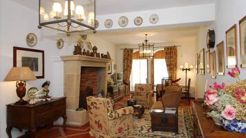 A traditional living room with a fireplace, floral armchairs, a wooden coffee table, and a variety of decorative items including a vase of flowers and framed pictures on the walls.