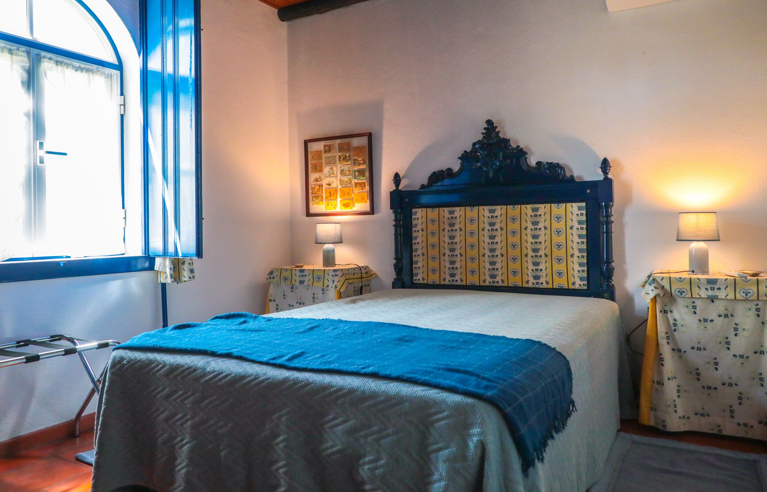 A bedroom with a bed, a window, a bedside table, a lamp, and a framed picture on the wall.