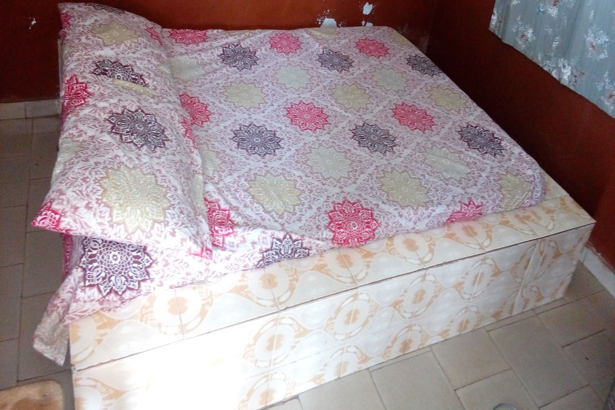 A bed with a patterned bedspread and pillows, a tiled floor, and a wall with a floral curtain.