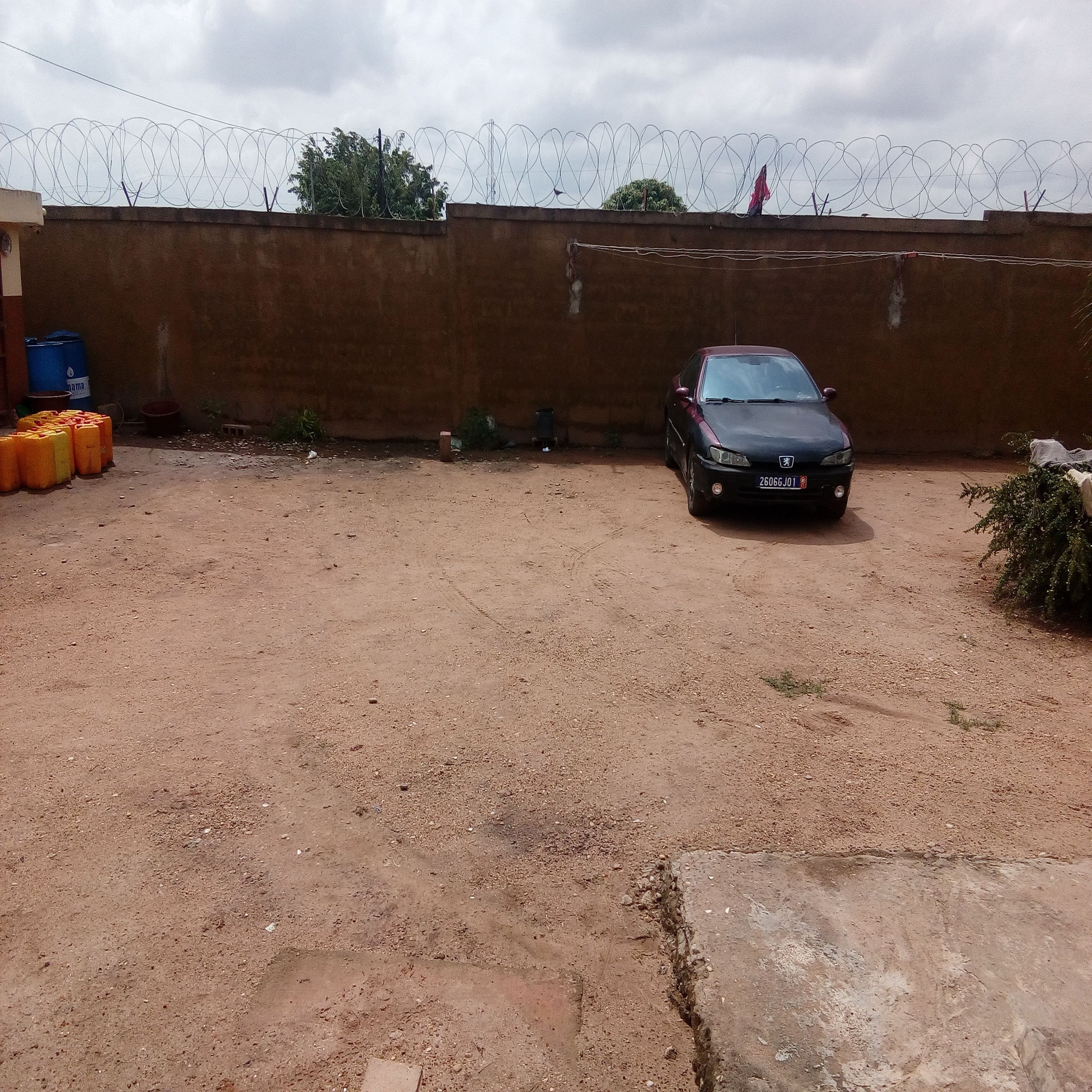 A parking area with a dirt ground, a car, and a wall topped with barbed wire.