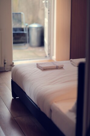 A neatly made bed with a folded towel, wooden flooring, and a view of the outdoors through an open door. - Camden Road Apartments (London)