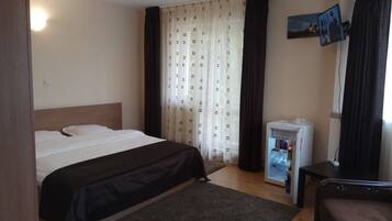 Habitación de hotel con cama, un refrigerador pequeño, televisor montado en la pared y una ventana con cortinas.