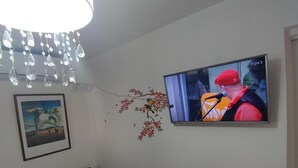 A living room with a television mounted on the wall, a decorative wall piece, a small table with items on it, and a patterned rug on the floor. - Eden's sweet place (Eilat)