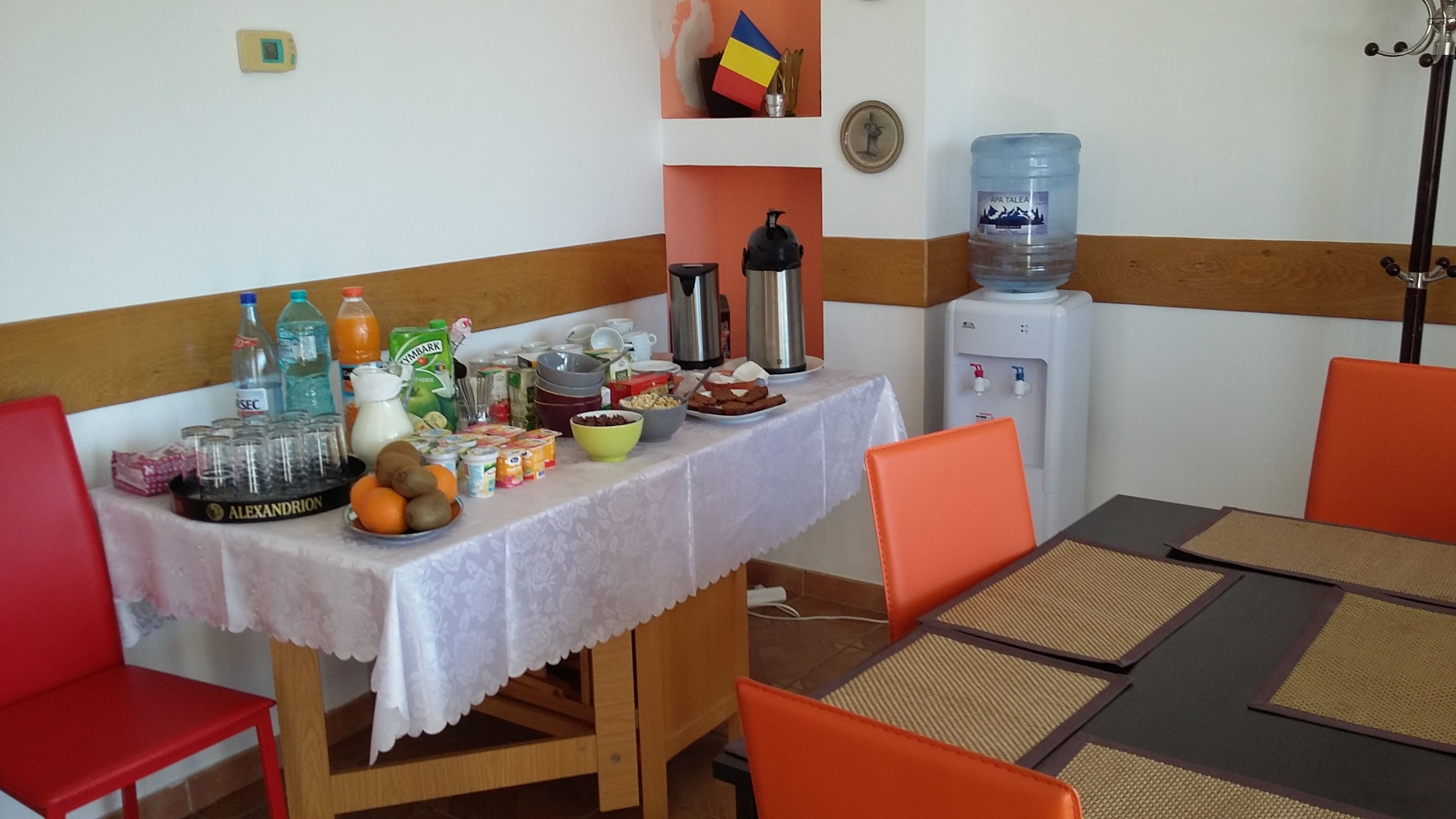 Ein Essbereich mit einem für vier Personen gedeckten Tisch, umgeben von orangefarbenen Stühlen. Auf dem Tisch befindet sich ein Wasserspender, eine Kaffeemaschine und verschiedene Speisen.