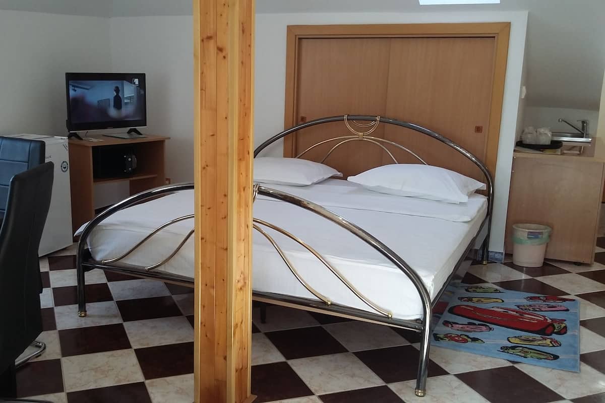 A room with a metal-framed bed, a desk with chairs, a television, and a checkered floor.