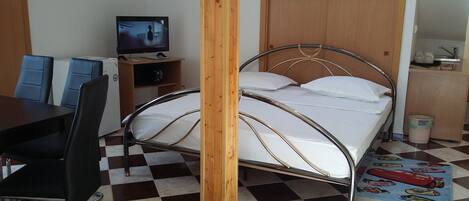 Quarto com cama de estrutura metálica, uma escrivaninha com cadeiras, televisão e piso xadrez.