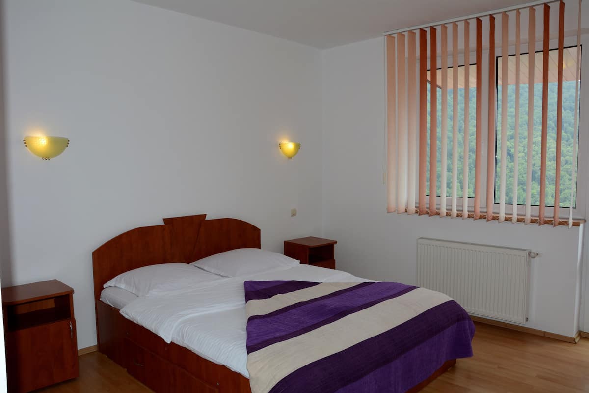 A bedroom with a wooden bed, two bedside tables, a window with vertical blinds, and two wall-mounted lights.