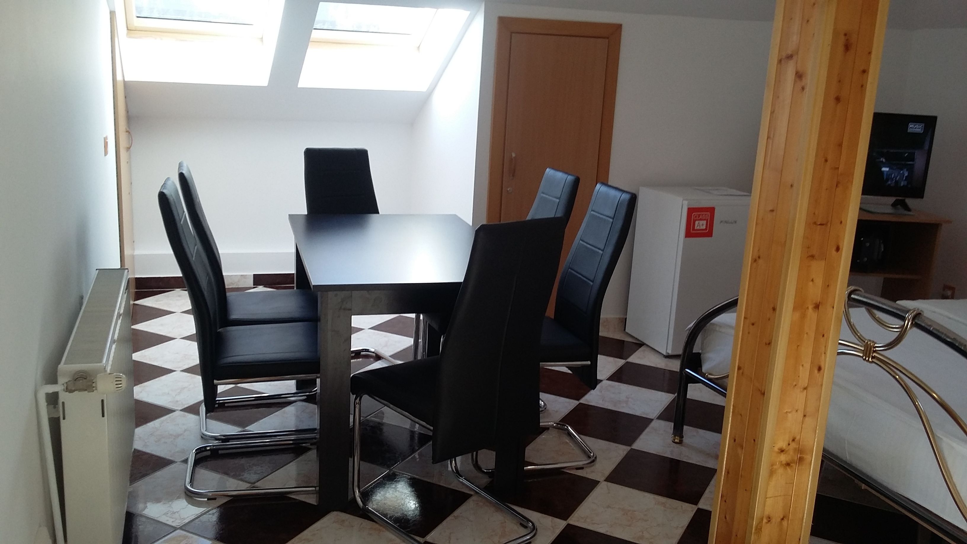 A room with a checkered floor, a table with four chairs, a bed, a television, and a window with skylight.