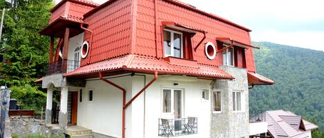 Ein zweistöckiges Haus mit rotem Dach, steinernen Fundament und einem Balkon mit Metallgeländer.