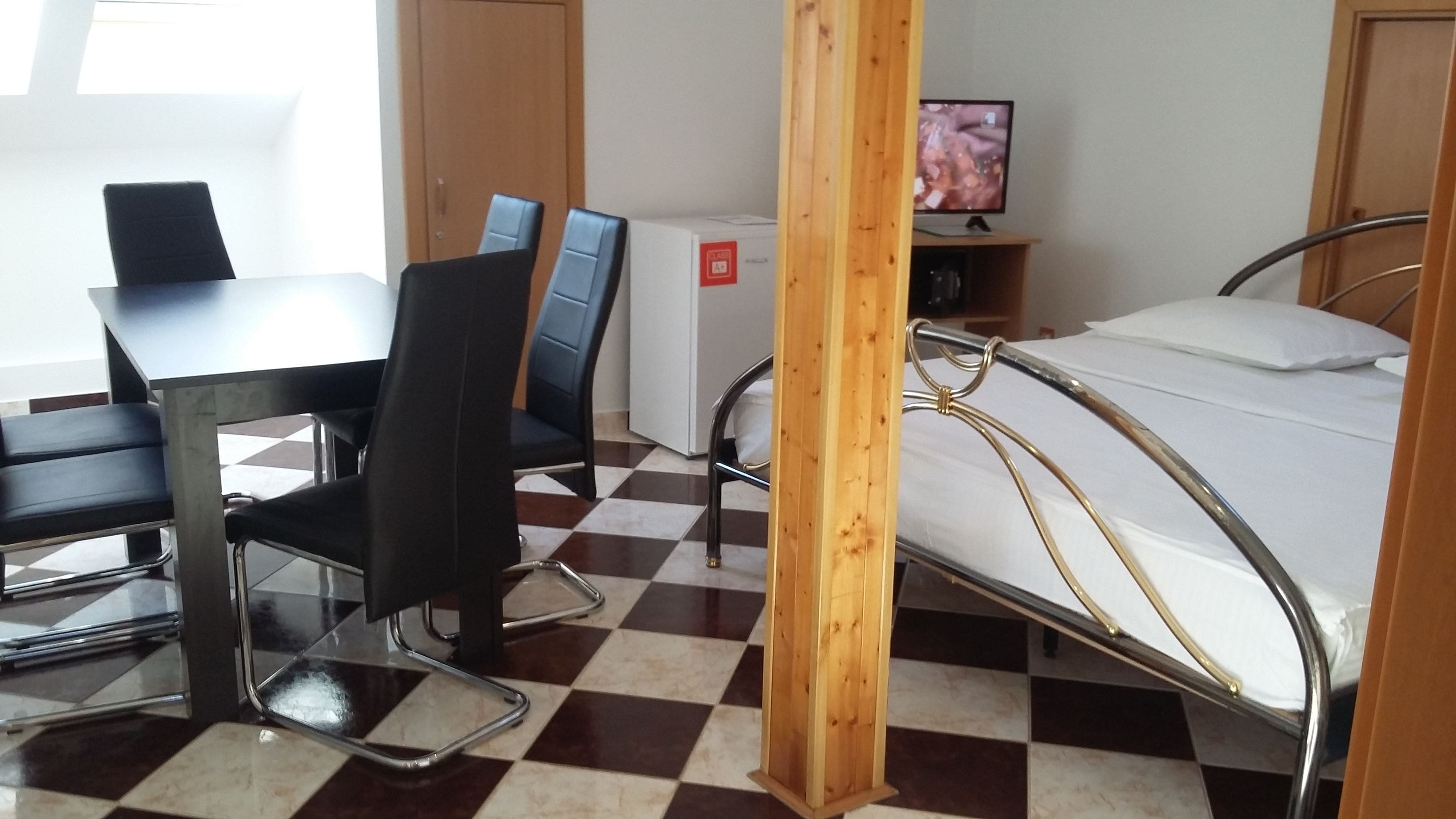A hotel room with a checkered floor, a bed with metal frame, a desk with chairs, and a television on a stand.