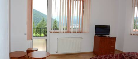 Une chambre à coucher avec un lit, un téléviseur posé sur une commode, une petite table en bois et une vue sur les montagnes par la fenêtre.