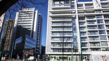 A modern multi-story building with a glass facade, multiple balconies, and a green entrance area.