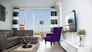 Een moderne woonkamer met een grijze bank, een glazen salontafel, een paarse fauteuil en een uitzicht op een stadsgezicht door grote ramen.