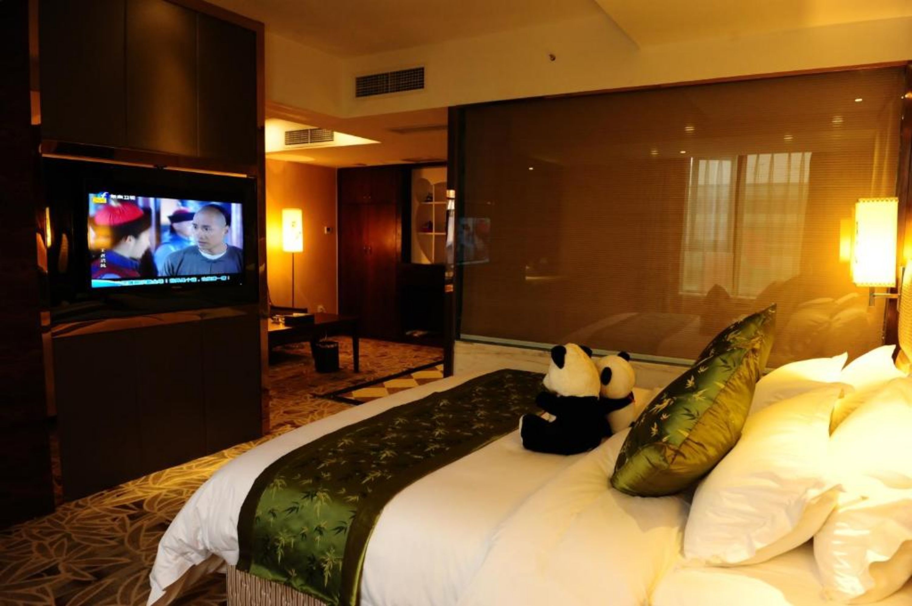 A hotel room with a bed, a television, a panda plush toy, and a large window.