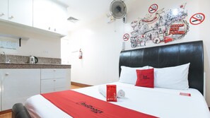 Une chambre d’hôtel avec un lit, une couvre-lit rouge et blanche, un oreiller orné du logo de l’hôtel, un ventilateur fixé au mur, un coin cuisine avec une bouilloire et un mur décoré d’œuvres d’art sur le thème du voyage.