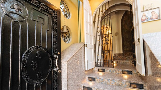 Una imponente entrada con una puerta negra ornamentada, una escalera de piedra con diseño de mosaico y una lámpara fijada a la pared.