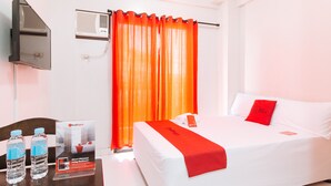 A hotel room with a bed, a TV, a window with orange curtains, and a table with water bottles and a RedDoorz sign. - RedDoorz near NAIA Terminal 1 (Parañaque)