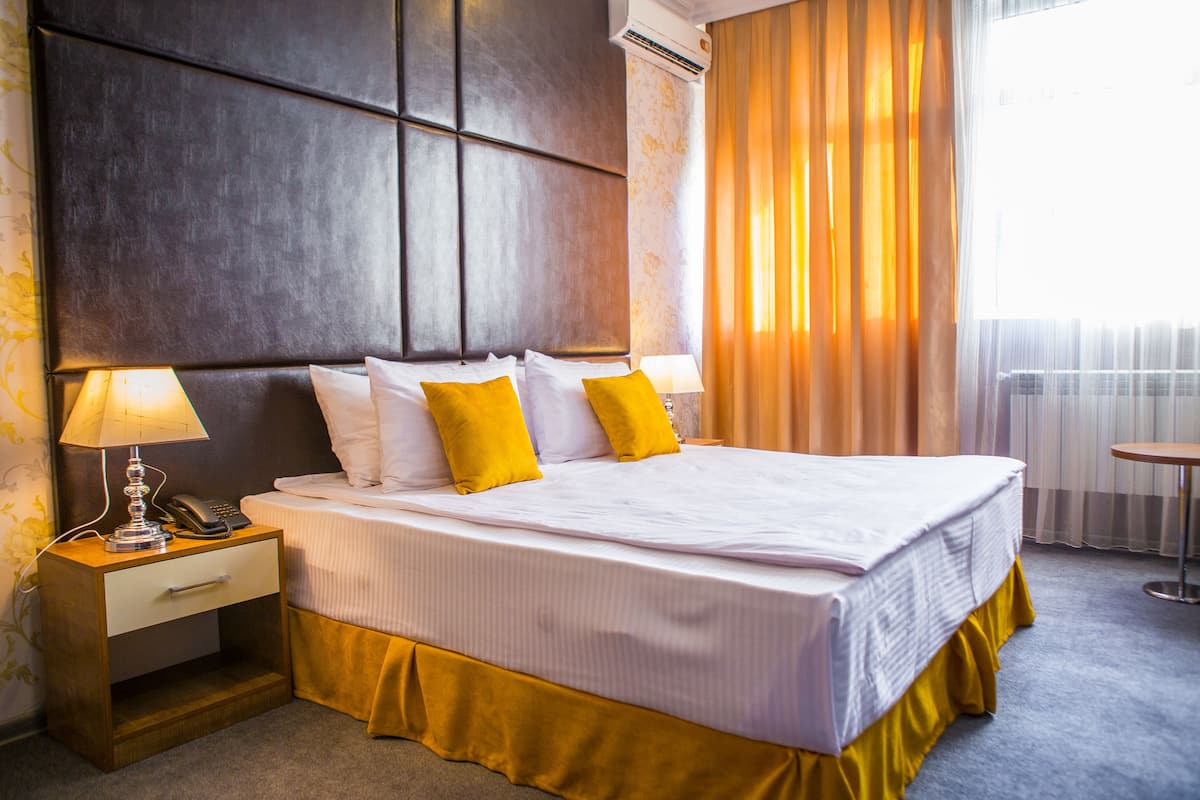 A hotel room with a large bed, two bedside tables with lamps, a small table, and a window with curtains.