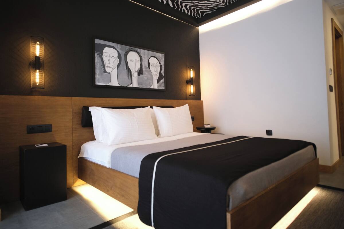 A modern hotel room with a large bed, a headboard with integrated lighting, a bedside table, and a wall-mounted artwork.