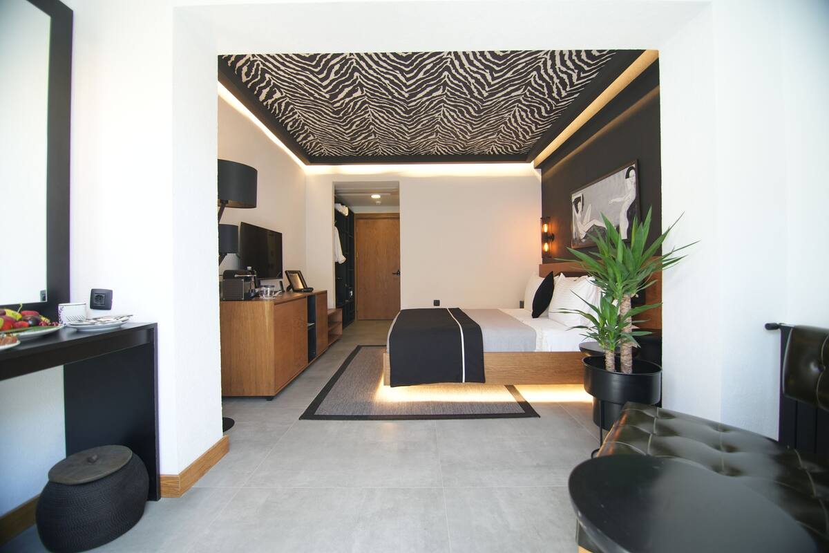 A modern hotel room with a zebra-patterned ceiling, a bed, a seating area, a kitchen, and a dining table.