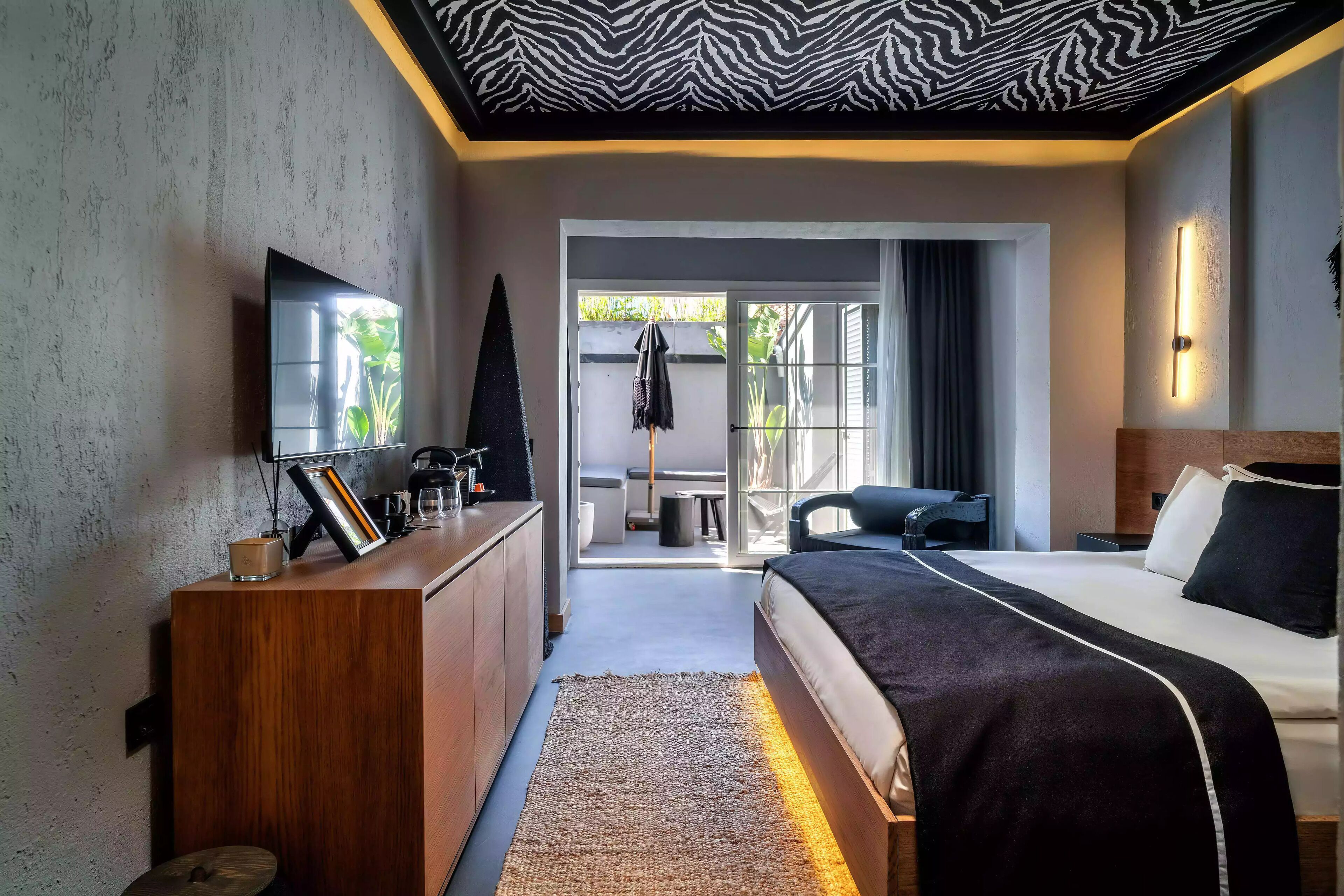A modern hotel room with a large bed, a wooden dresser, a flat-screen TV, and a view of the outdoors.