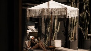 A person relaxing on a lounge chair under a fringed umbrella, with a pool and cacti in the background. - The Local House Alaçatı (Cesme)