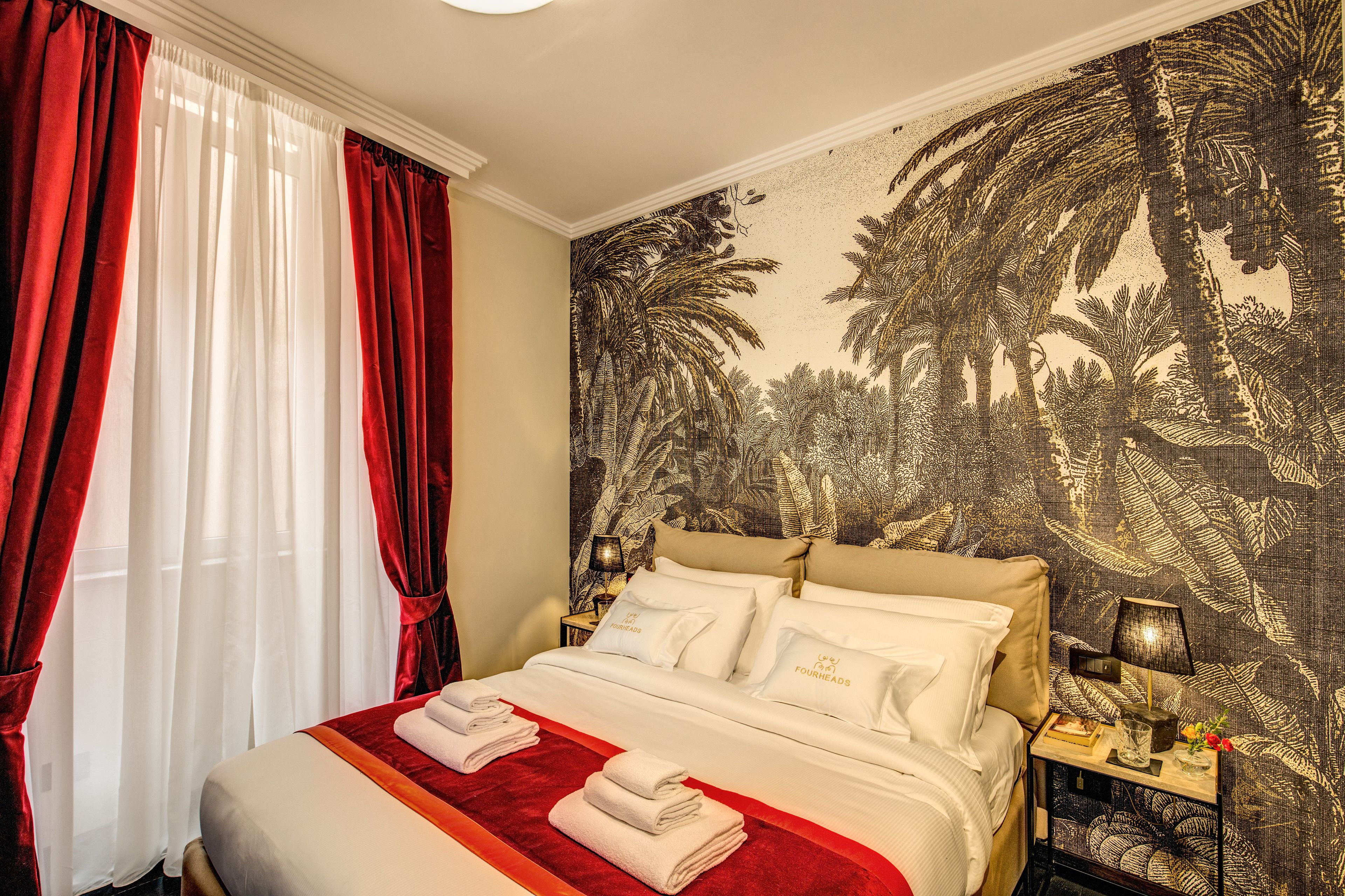 Une chambre d’hôtel avec un grand lit, des rideaux rouges, un papier peint aux motifs tropicaux et une table de chevet avec une lampe et un livre.