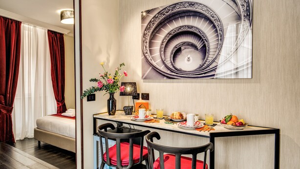 A hotel room with a bed, a dining area with a table set for breakfast, and a large spiral staircase artwork on the wall. - PAX Hotel (Rome)