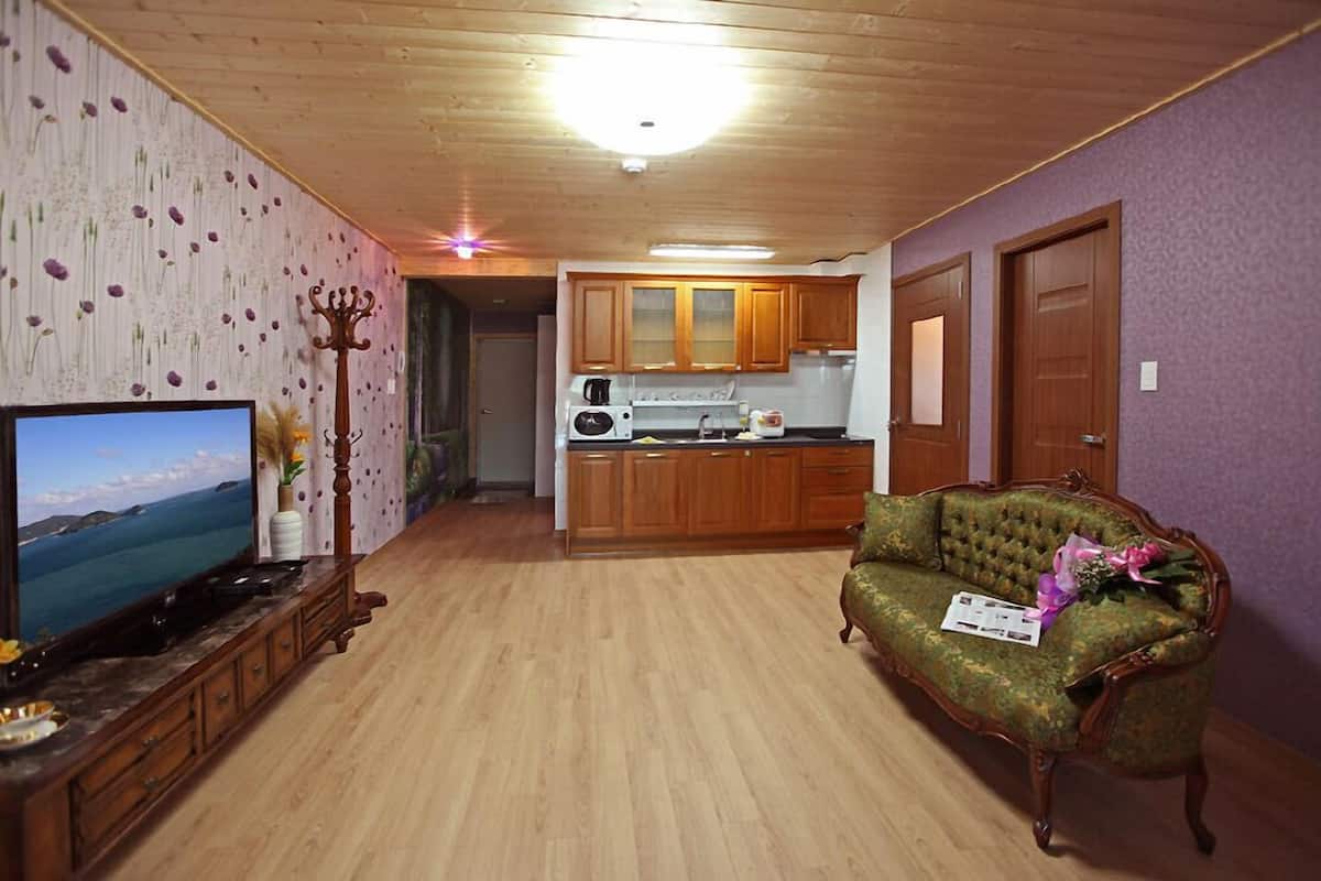 Sala de estar con televisor, sofá verde y mesa de centro de madera. Se observa una zona de cocina con armarios de madera y un microondas.