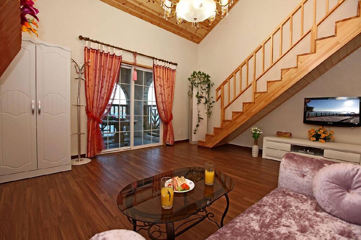 Amplia sala de estar con escalera de madera, una mesa de centro de vidrio con comida y bebidas, un sofá y un televisor sobre un mueble blanco.