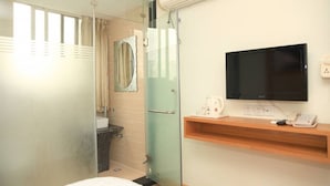 Quarto de hotel com box de vidro, televisão montada na parede e prateleira de madeira com telefone e uma pequena mesa.