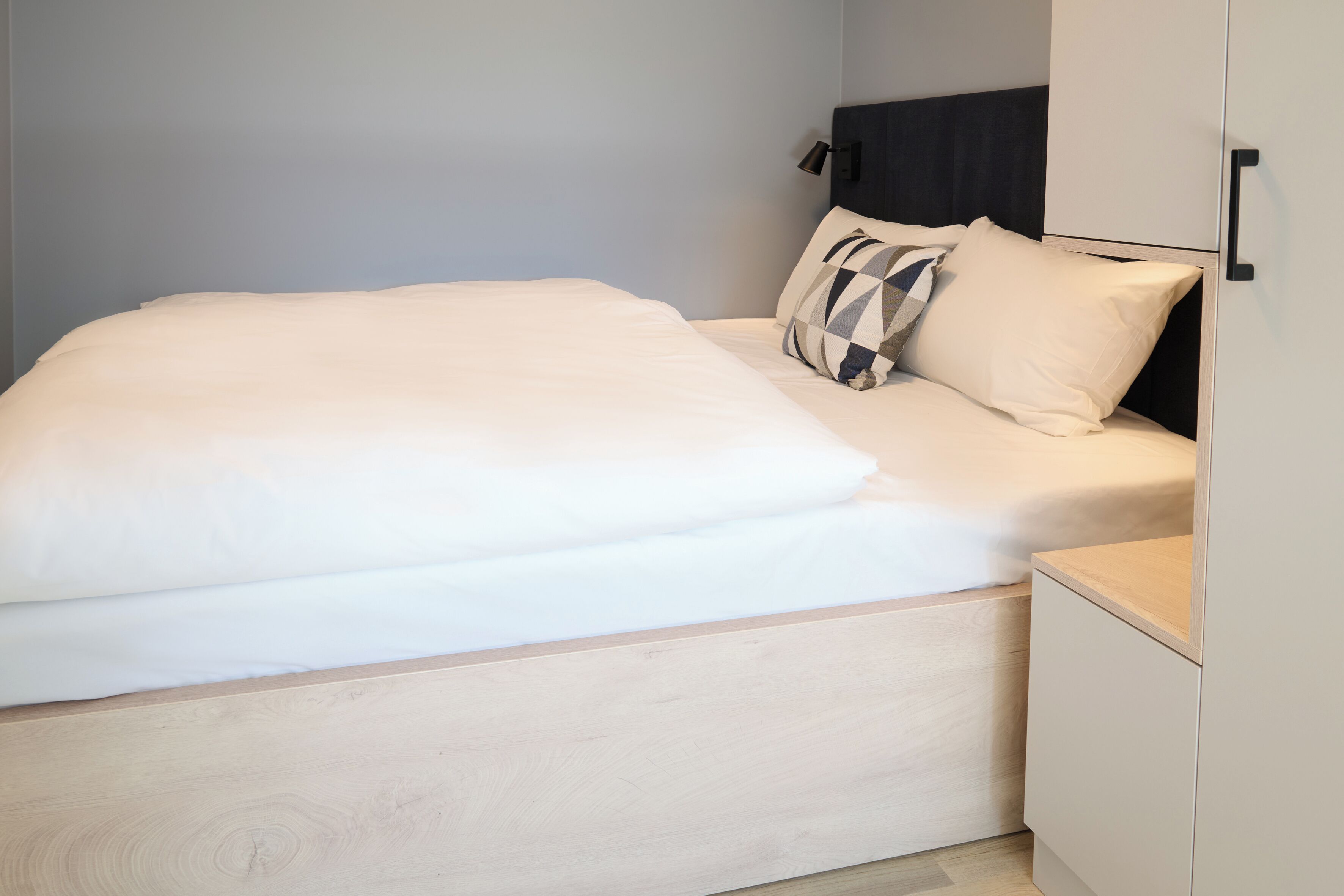 En pænt redt seng med hvide sengetøj og pude med mønster, et natbord og en garderobe med lyse trædetaljer.