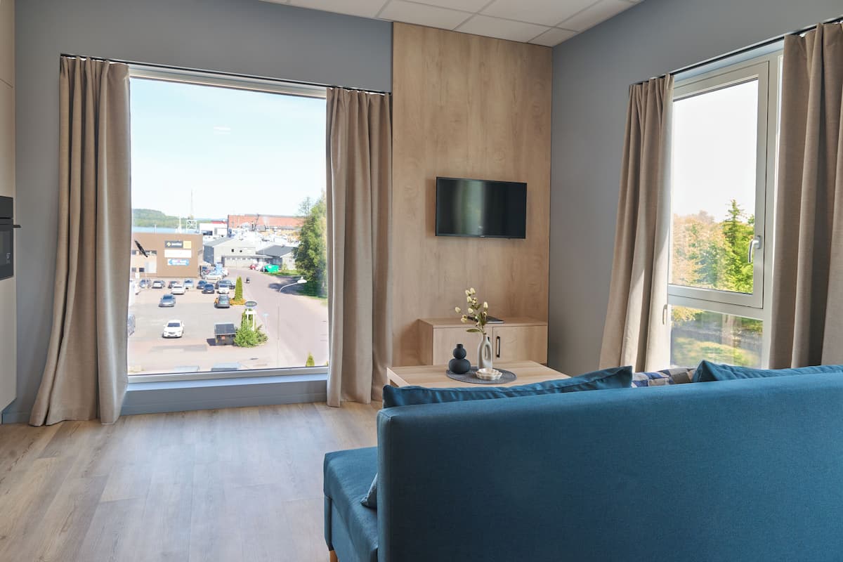 Een hotelkamer met een groot raam, een flatscreen televisie aan de muur, een houten kast en een blauwe bank.
