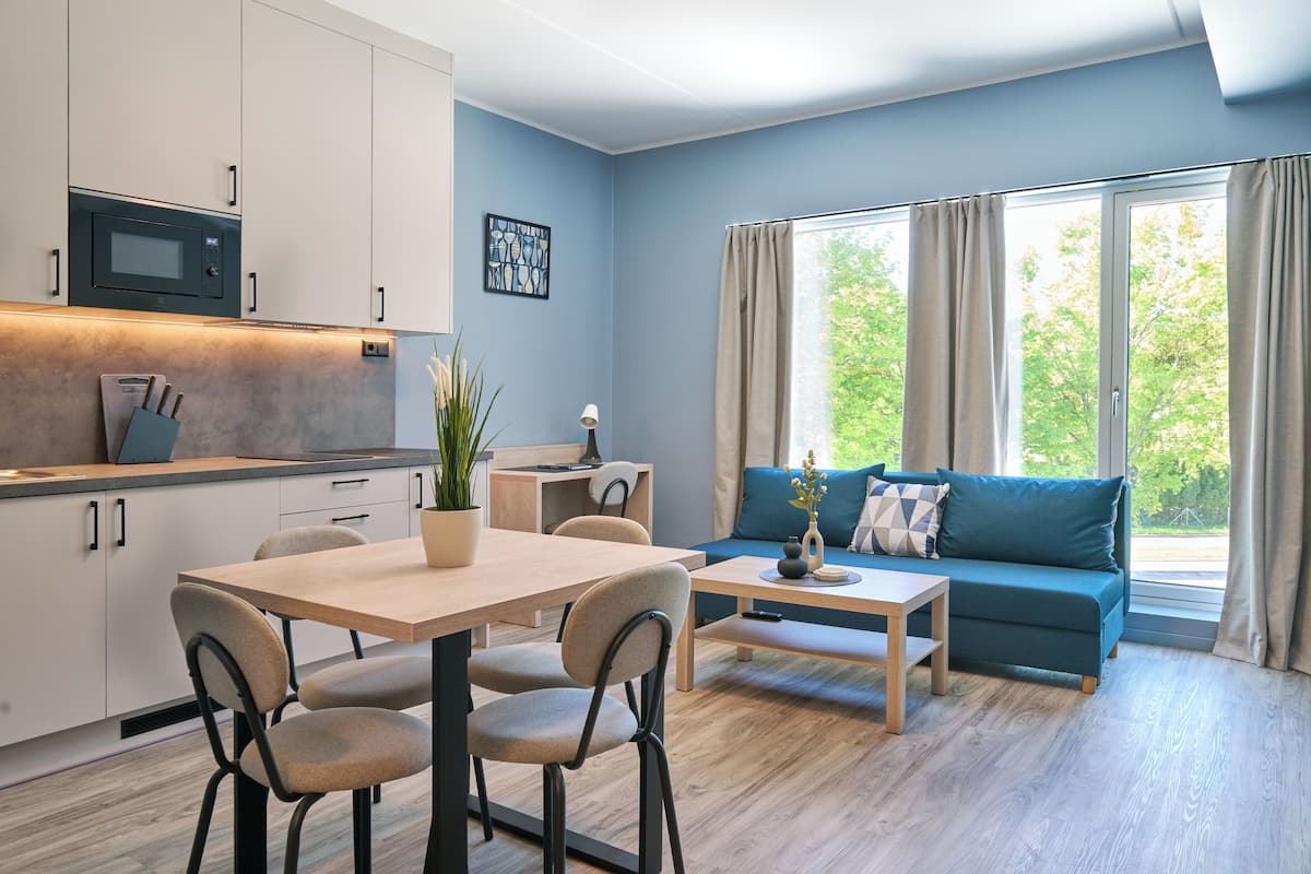 Een moderne keuken met witte kasten, een magnetron, en een eethoek met een tafel en stoelen. Er is een blauwe bank en een raam met gordijnen.
