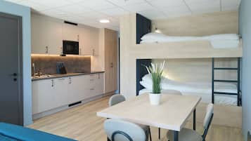 A compact hotel room with a bunk bed, a kitchenette, a dining area with a table and chairs, and a potted plant.