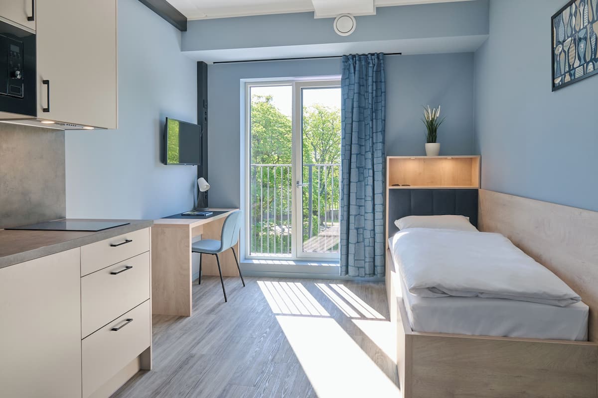 Een moderne slaapkamer met een bed, een bureau en een computer. Er is een balkon met uitzicht op bomen.