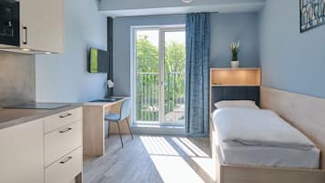 Un dormitorio moderno con cama, escritorio y computadora. Hay un balcón con vista a árboles.