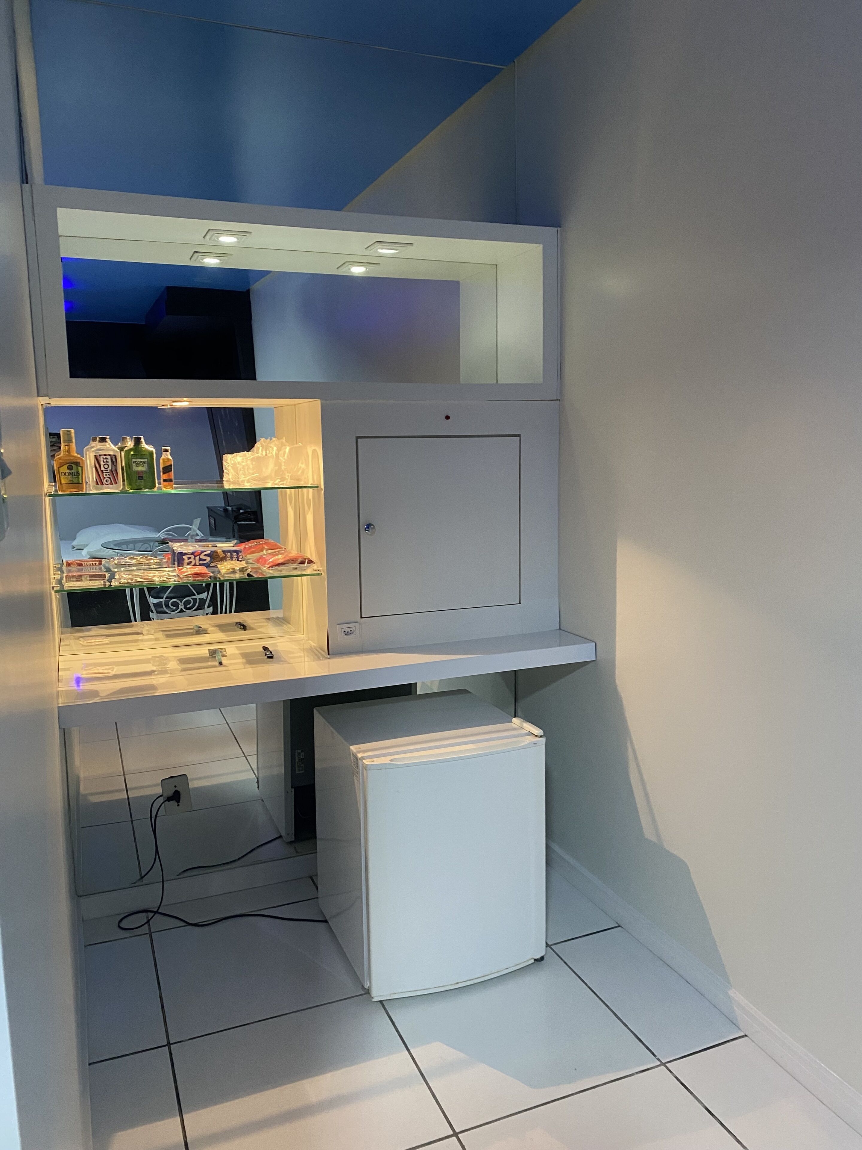 Una kitchenette compacta con refrigerador, microondas y repisas con diversos artículos.