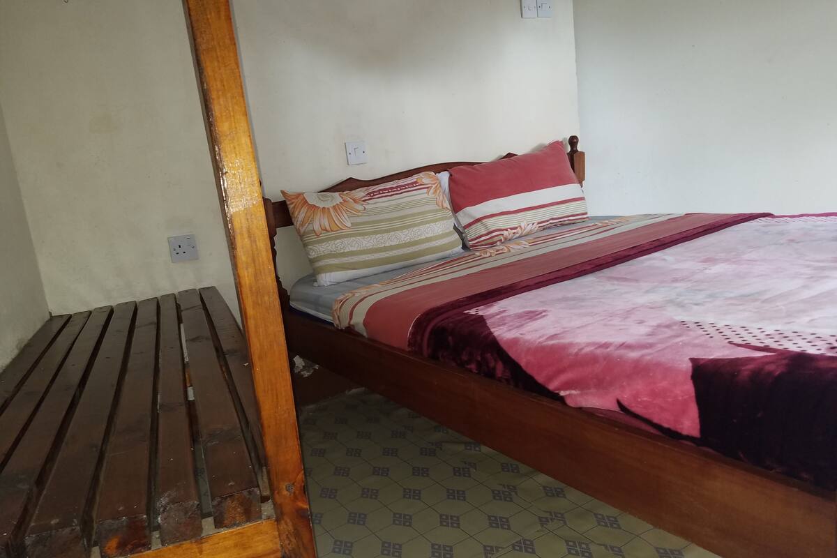 Een enkel bed met houten frame, een bed met een decoratief sprei en kussens met bloemmotieven.