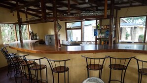 A bar area with a wooden counter, metal chairs, and a view of the outside through large windows.