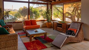Un salon lumineux, avec un canapé, un fauteuil et une table basse, de grandes fenêtres offrant une vue sur la verdure et les collines, et une plante en pot.