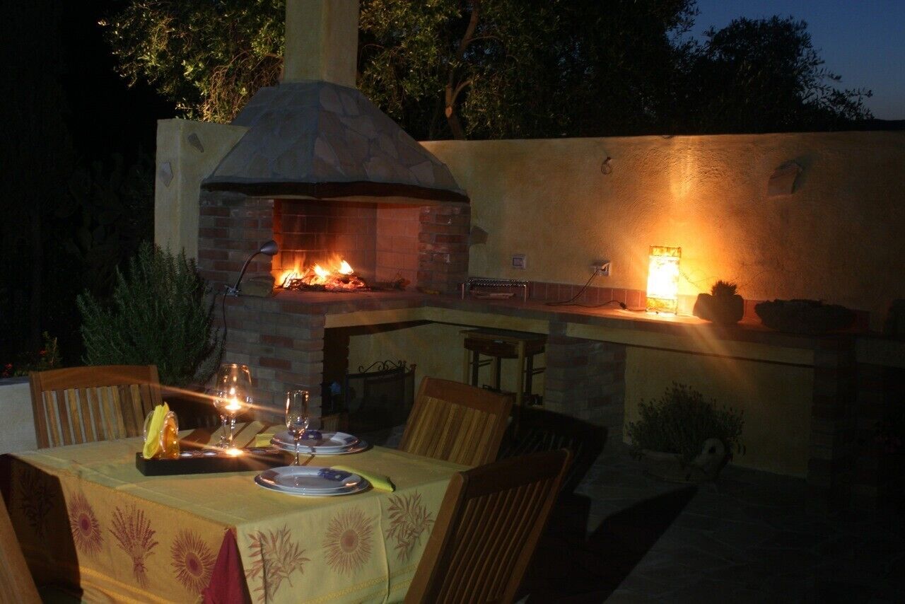 An outdoor dining area with a brick oven and grill, surrounded by chairs and a table set for two.