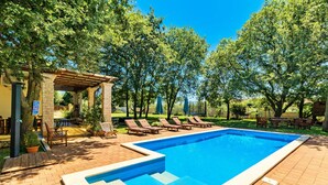 Une piscine rectangulaire à l’eau d’un bleu limpide, entourée d’une terrasse en briques. On y trouve des chaises longues et une pergola avec un toit, ainsi que des arbres en arrière-plan.