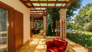 Un patio couvert avec un sol en pierre, un fauteuil en cuir rouge et une vue sur une piscine et un jardin.