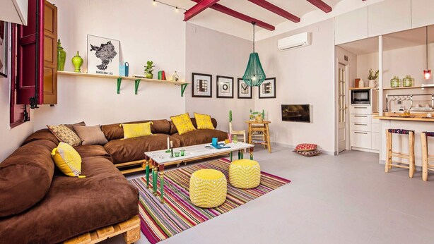 A cozy living room with a brown sofa, colorful cushions, a striped rug, and a kitchen area with white cabinets and a wooden bar stool.