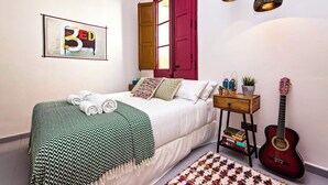 A bedroom with a bed, pillows, a guitar, a guitar stand, a bedside table with books, and a wall with a framed picture.